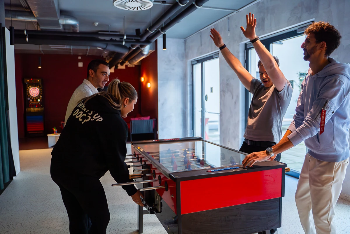 Vier junge Erwachsene spielen Tischfußball in einem modernen Raum. Spaß und Teamwork im Mittelpunkt.